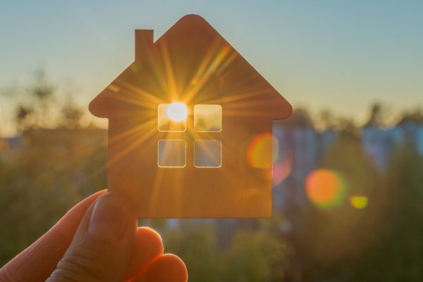 Small cardboard cutout of a house held to the sky at sunset with a sunburst peeking through its window.